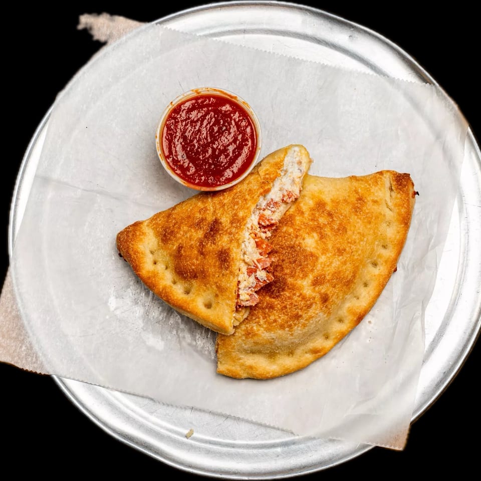 A golden-brown calzone, sliced open to reveal a cheesy filling, served with a side of marinara sauce.