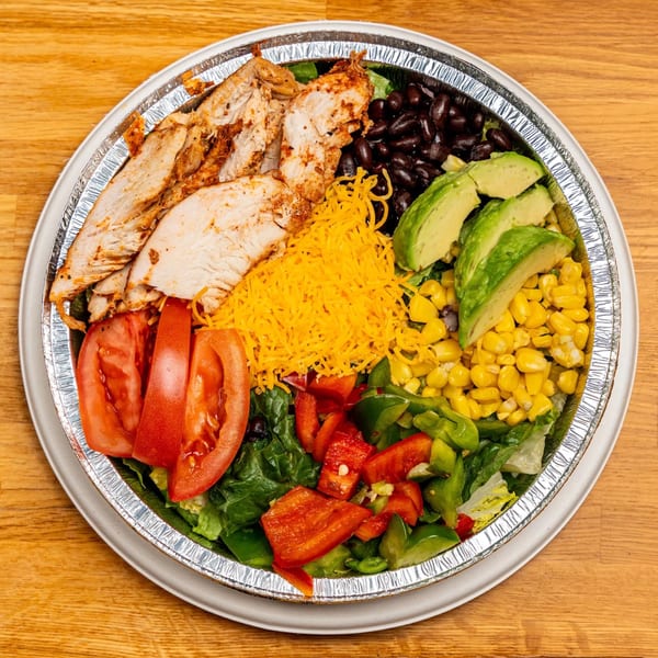 Chicken salad with black beans, avocado, corn, tomatoes, bell peppers, and shredded cheese in a foil container.