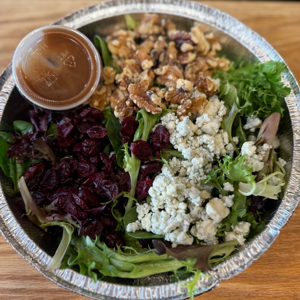 A salad in a foil container with mixed greens, walnuts, dried cranberries, blue cheese, and dressing.
